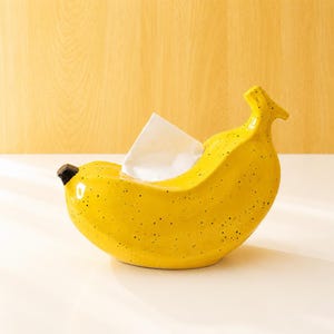 Op de afbeelding: Een felgele bananenvormige tissuehouder met een glanzende afwerking en kleine zwarte spikkels. Het object heeft een donkerbruine punt aan het ene uiteinde en een steeltje aan het andere, met een witte tissue die uit de opening steekt.
