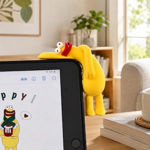 May include: A yellow, cartoon-style character figurine with large white eyes and a red mouth, draped over the top edge of a black tablet screen. The tablet displays a digital drawing of the same yellow character with a green scarf and a red heart.