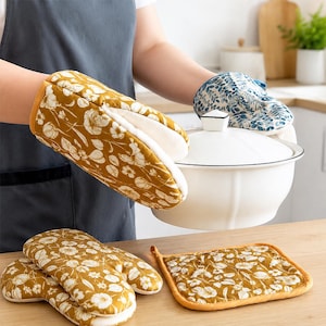 Può includere: Un set di tessili da cucina che include due guanti da forno color giallo senape con motivo floreale bianco, un guanto da forno con motivo a foglie blu e bianco e una presina quadrata color giallo senape, esposti con una pentola in ceramica.