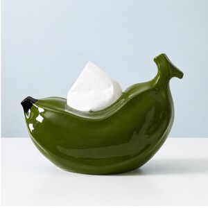 Puede incluir: Un soporte para pañuelos de cerámica en color verde oliva brillante con forma de plátano curvado. El objeto presenta una punta marrón oscuro en un extremo y un tallo en el otro, con un pañuelo blanco asomando por la abertura central.
