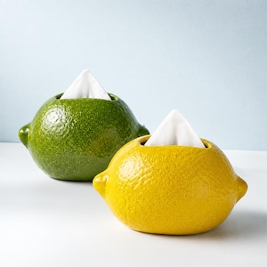 Puede incluir: Dos porta pañuelos de cerámica con forma de cítricos. Uno es de color verde texturizado, parecido a una lima, y el otro es de color amarillo texturizado, parecido a un limón. Cada recipiente tiene una abertura superior con un pañuelo blanco.