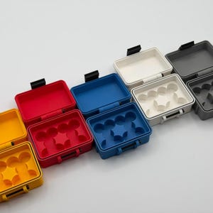 May include: A collection of rectangular plastic storage cases in yellow, red, blue, white, and grey. Each case features a hinged lid with a black latch and an interior insert with multiple circular and star-shaped compartments for organising small items.