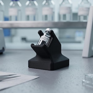 May include: A black, angled laboratory vial holder made of a matte material, securely cradling a small glass vial with a white label that reads SAMPLE VIAL-04 08/2024. The holder features a sturdy, wide base and a slanted top opening.