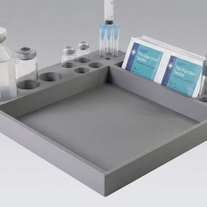 May include: A grey square organiser tray with multiple circular cutouts for medical vials and syringes. The tray holds a large clear glass vial, several smaller glass vials, a syringe, and two white packets labelled Pre-injection Swabs.
