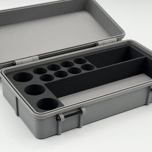 May include: A rectangular grey storage case with a hinged lid and a black foam insert. The interior features ten circular cutouts for small cylindrical items and two larger rectangular compartments for organised storage of various small objects.