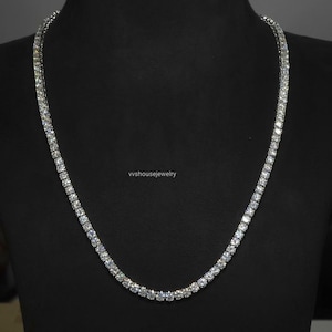 May include: A silver-toned tennis necklace featuring a continuous row of round, clear, faceted stones set in a metallic frame. The necklace is displayed on a black bust, and the text vvhousejewelry is visible in the center of the image.