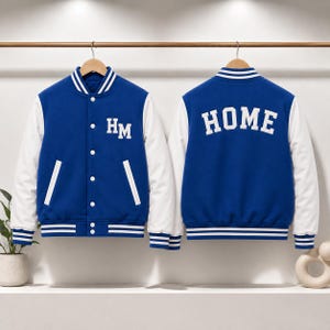 Op de afbeelding: Een blauw met wit varsity jack, getoond van de voor- en achterkant. De voorkant is voorzien van de letters HM in het wit, terwijl de achterkant het woord HOME in grote witte letters toont. Het jack heeft witte mouwen, een gestreepte kraag en boorden.