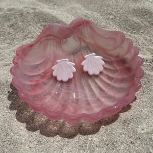 Puede incluir: Un par de pendientes de botón en forma de concha de color rosa claro que descansan dentro de un plato de vidrio translúcido, jaspeado en rosa y blanco, con forma de concha de vieira. El plato presenta un borde festoneado y un acabado brillante.