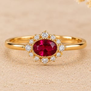 Op de afbeelding: Een gouden ring met een centrale ovaal geslepen dieprode edelsteen, omringd door een halo van kleine, ronde, helderwitte steentjes. De zetting is gedetailleerd met een geschulpte rand en de ringband is van glad, gepolijst goudkleurig metaal.