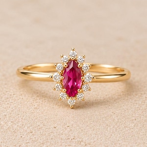 May include: A gold ring featuring a central marquise-cut vibrant pink gemstone surrounded by a halo of twelve small round white stones. The band is smooth and polished, showcasing a delicate and symmetrical design with a bright metallic finish.