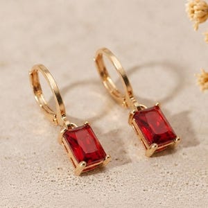 Peut inclure: Une paire de boucles d'oreilles créoles dorées avec des pierres précieuses rectangulaires rouge foncé suspendues à chaque anneau. Les boucles d'oreilles présentent une finition métallique polie et un mécanisme de fermeture sécurisé.