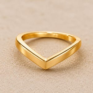 Pode incluir: Um anel de ouro polido com um design distinto em forma de V. A banda tem uma superfície lisa e sólida com um acabamento metálico reflexivo, formando uma ponta afiada no centro da silhueta do anel, exibindo uma estrutura limpa e simples.