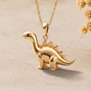 Könnte beinhalten: Eine goldfarbene Halskette mit einem Anhänger in Form eines Stegosaurus-Dinosauriers. Der Anhänger hat eine glatte, polierte Oberfläche mit einem kleinen schwarzen Auge und einer Reihe dreieckiger Platten auf dem Rücken.