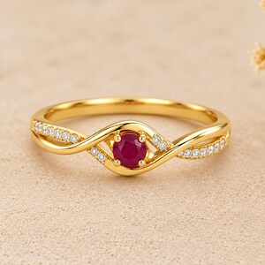 May include: A gold ring featuring a central round-cut red ruby gemstone held in a four-prong setting. The band has a twisted, crossover design adorned with small, clear pavé-set diamonds along both sides of the shank, creating a delicate look.