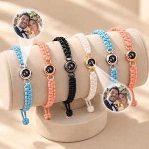 Può includere: Sei braccialetti in corda intrecciata nei colori blu, pesca, nero, bianco, azzurro e pesca, ciascuno con un ciondolo centrale in metallo circolare contenente una piccola proiezione di una foto di una coppia sorridente.