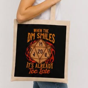 Op de afbeelding: Een beige canvas draagtas met een zwarte vierkante opdruk van een twintigzijdige dobbelsteen omringd door oranje en rode vlammen. De tekst luidt: WHEN THE DM SMILES IT'S ALREADY Too Late. De tas heeft twee schouderbanden en een rechthoekige vorm.