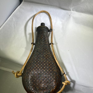 May include: An antique copper powder flask with a leather carrying strap. The flask has a decorative embossed pattern and a small spout.