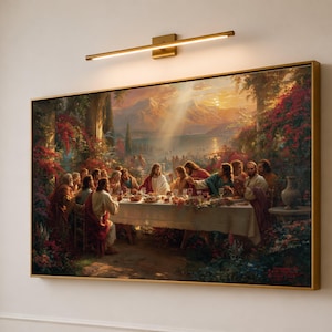 May include: A rectangular framed wall art print depicting a group of people gathered around a long table for a meal in an outdoor garden setting with mountains in the background, illuminated by a gold-toned horizontal wall light fixture above.