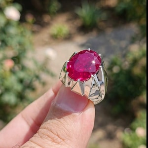 Puede incluir: Un anillo de plata con una gran piedra preciosa de color rojo vibrante y corte ovalado, engastada en una montura metálica de perfil alto con múltiples garras. La banda es de plata pulida y la piedra muestra un tono carmesí saturado.