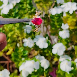 Op de afbeelding: Een gouden ring met een centrale vliegervormige rode edelsteen, geflankeerd door kleine heldere steentjes aan weerszijden. De ring wordt vastgehouden door een metalen pincet, wat het fijne metaalwerk en de levendige rode kleur benadrukt.