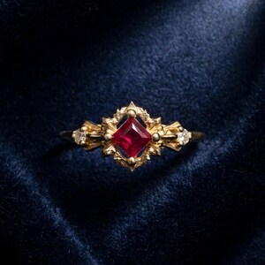 May include: A gold ring featuring a central square-cut red gemstone set within an ornate, textured metal frame. The band includes small, clear accent stones on either side of the central setting, showcasing intricate metalwork details.