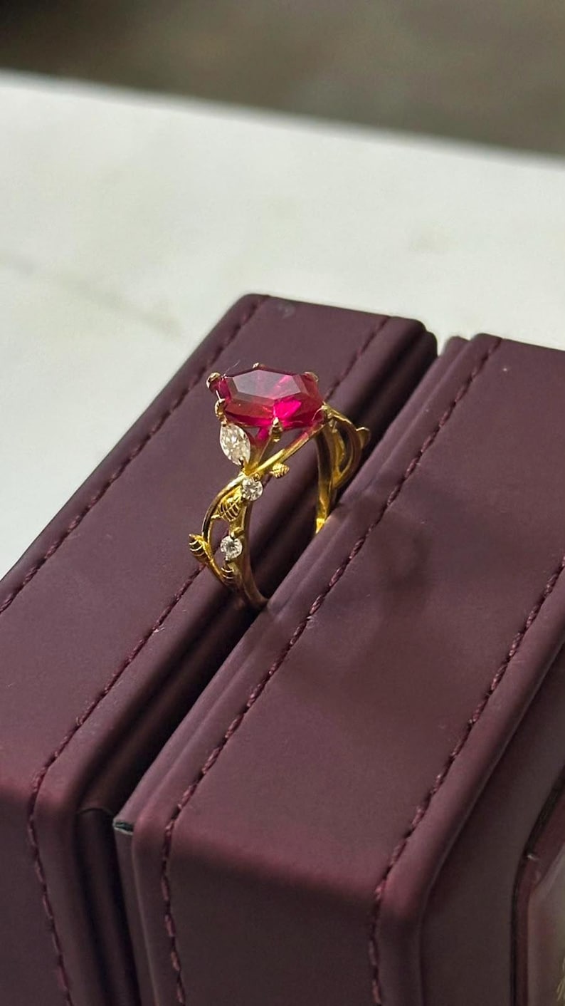 Op de afbeelding: Een gouden ring met een grote, rechthoekige, dieprode edelsteen als hoofdsteen. De ringband is ontworpen met een rankachtig motief, waarin kleine, heldere, rondgeslepen accentstenen en &eacute;&eacute;n markiesgeslepen steen zijn verwerkt.