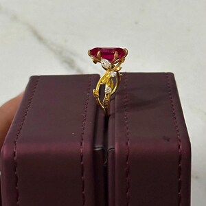Op de afbeelding: Een gouden ring met een grote, ovaal geslepen dieprode edelsteen op een band met delicaat, rankachtig metaalwerk en kleine, heldere accentstenen. De ring staat verticaal op de rand van een donkerpaarse rechthoekige doos.