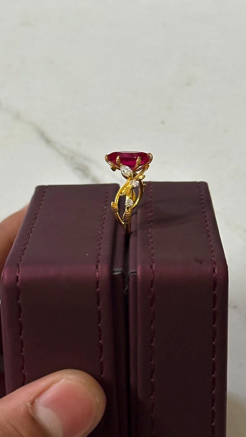 Op de afbeelding: Een gouden ring met een grote, ovaal geslepen dieprode edelsteen op een band met delicaat, rankachtig metaalwerk en kleine, heldere accentstenen. De ring staat verticaal op de rand van een donkerpaarse rechthoekige doos.