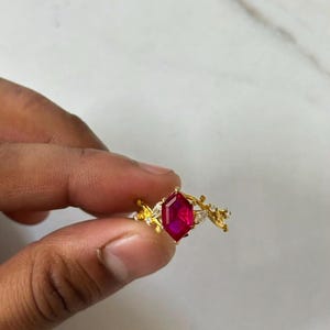 Op de afbeelding: Een gouden ring met een centrale zeshoekig geslepen, levendig rode edelsteen in een klauwzetting. De ringband is ontworpen met een gestructureerd, takachtig motief en is aan weerszijden van de middelste steen bezet met kleine, heldere markiesstenen.