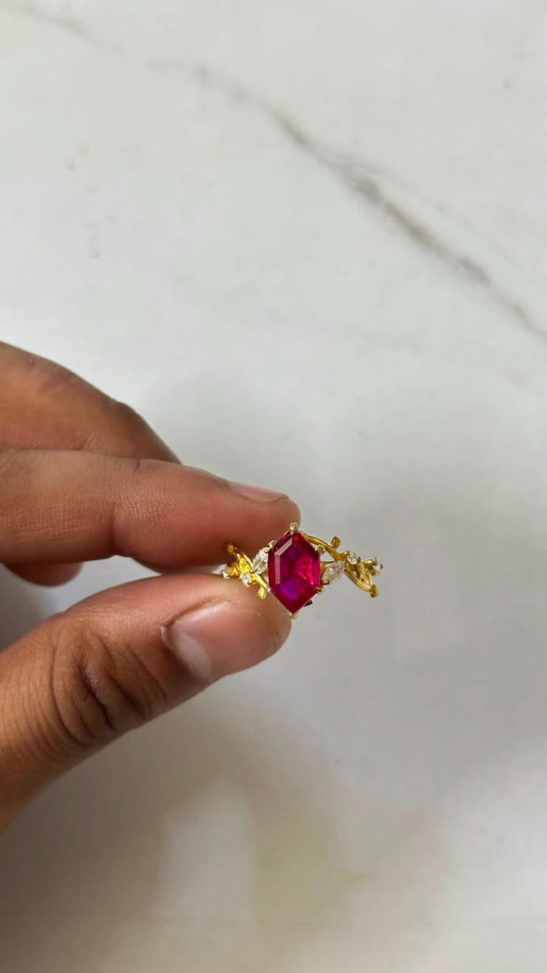 Op de afbeelding: Een gouden ring met een centrale zeshoekig geslepen, levendig rode edelsteen in een klauwzetting. De ringband is ontworpen met een gestructureerd, takachtig motief en is aan weerszijden van de middelste steen bezet met kleine, heldere markiesstenen.