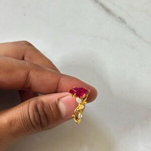 Op de afbeelding: Een gouden ring met een opvallende, marquise-geslepen rode edelsteen in het midden, geaccentueerd door kleine heldere steentjes langs een rankachtige band. Het metaal heeft een gepolijste gele afwerking met gedetailleerde versieringen.