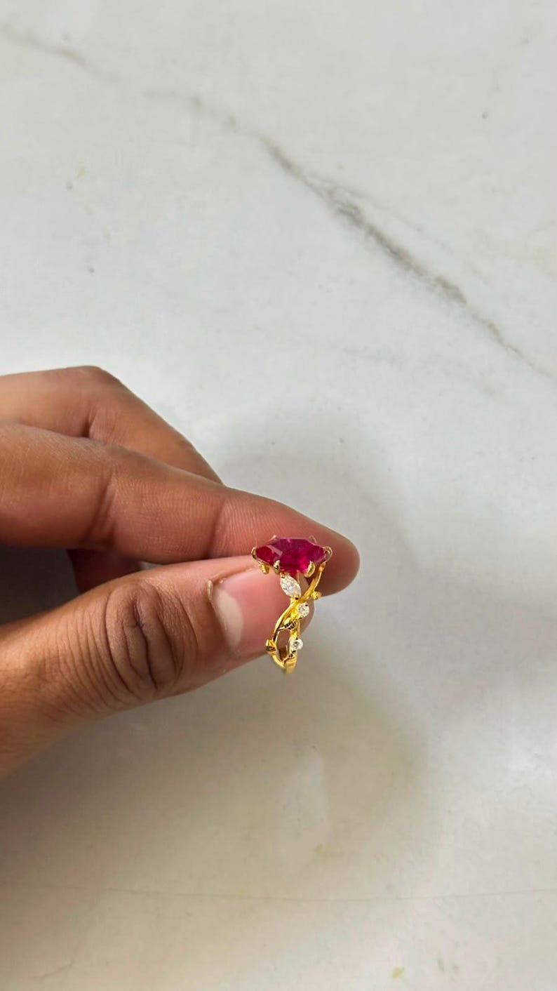 Op de afbeelding: Een gouden ring met een opvallende, marquise-geslepen rode edelsteen in het midden, geaccentueerd door kleine heldere steentjes langs een rankachtige band. Het metaal heeft een gepolijste gele afwerking met gedetailleerde versieringen.