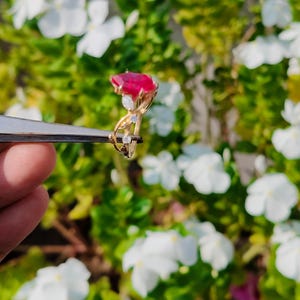 Op de afbeelding: Een gouden ring met een grote, gefacetteerde, rechthoekige rode edelsteen als middelpunt, geaccentueerd door drie kleine, heldere, rondgeslepen stenen die langs de delicate, gedraaide gouden band naar de zetting toe zijn geplaatst.