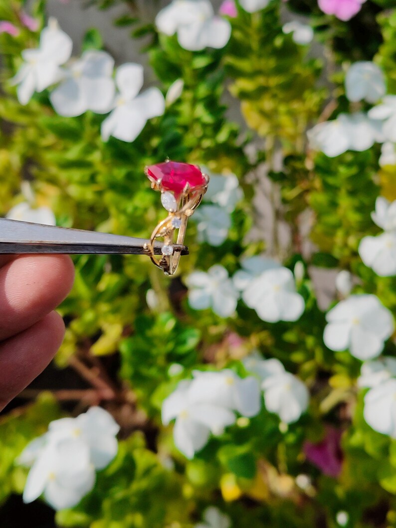 Op de afbeelding: Een gouden ring met een grote, gefacetteerde, rechthoekige rode edelsteen als middelpunt, geaccentueerd door drie kleine, heldere, rondgeslepen stenen die langs de delicate, gedraaide gouden band naar de zetting toe zijn geplaatst.
