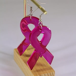 Peut inclure: Une paire de boucles d'oreilles en forme de ruban de sensibilisation, réalisées en acrylique rose vif translucide. Chaque pendentif en forme de ruban est suspendu à un crochet d'oreille en métal argenté sur un présentoir doré.