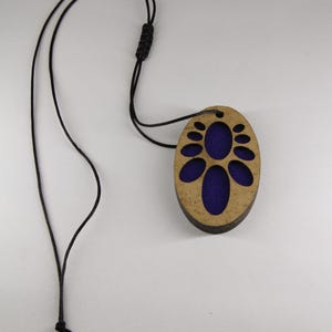 Peut inclure: Un collier pendentif de forme ovale avec un cadre en bois brun clair et un motif découpé révélant une incrustation en tissu violet foncé. Le pendentif est suspendu à un cordon noir fin doté d'un nœud coulissant réglable en longueur.