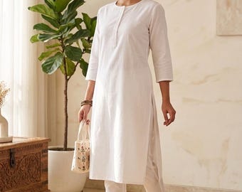 Può includere: Una tunica bianca a maniche lunghe con scollo rotondo e abbottonatura parziale, abbinata a pantaloni bianchi a gamba dritta coordinati. L'outfit è completato da una piccola borsa di colore chiaro con un delicato motivo floreale.