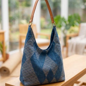Peut inclure: Un sac à bandoulière de style hobo en tissu bleu orné d'un motif répétitif de vagues en forme d'éventail blanc. Le sac présente un design structuré en patchwork de losanges et une bandoulière en cuir beige fixée par des anneaux dorés.