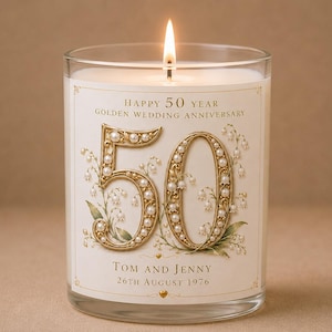 Op de afbeelding: Een kaars in helder glas met witte was en een brandende lont. Het etiket bevat de tekst "HAPPY 50 YEAR GOLDEN WEDDING ANNIVERSARY", "TOM AND JENNY" en "26TH AUGUST 1976" naast een groot goudkleurig getal 50 versierd met witte parels.