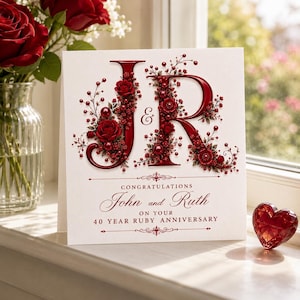 May include: A white greeting card featuring the large red letters J and R decorated with intricate red roses and small beads. The text reads: CONGRATULATIONS John and Ruth ON YOUR 40 YEAR RUBY ANNIVERSARY. A red heart-shaped crystal sits nearby.
