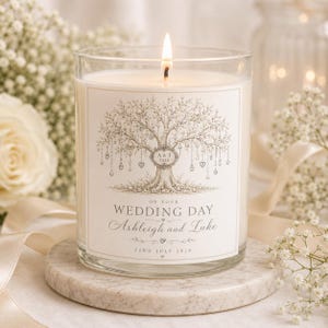 May include: A clear glass candle with a lit wick, featuring a label with a detailed illustration of a tree adorned with hanging ornaments. The text reads: ON YOUR WEDDING DAY, Ashleigh and Luke, 22ND JULY 2026, and A&T 2026 within a central frame.