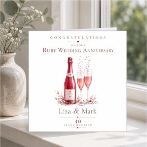 Op de afbeelding: Een vierkante wenskaart met de tekst CONGRATULATIONS ON YOUR RUBY WEDDING ANNIVERSARY, Lisa & Mark, en 40 YEARS MARRIED. Het ontwerp bevat een aquarelillustratie van een rode champagnefles en twee glazen met bubbels.