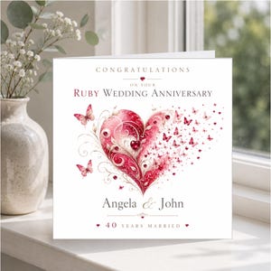 May include: A square greeting card with white background featuring the text CONGRATULATIONS ON YOUR RUBY WEDDING ANNIVERSARY. A large red heart design with swirling patterns and small butterflies is centered. Below, the names Angela & John appear with 40 YEARS MARRIED.