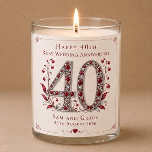 Può includere: Una candela in vetro trasparente con cera bianca e stoppino acceso. L'etichetta riporta il testo Happy 40th Ruby Wedding Anniversary, il numero 40 con un design di gioielli rossi e argento, e i nomi Sam and Grace con la data 25th August 2026.