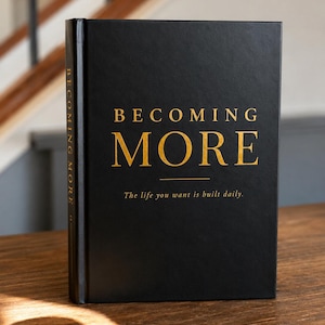 May include: A black hardcover book with gold foil lettering on the front cover. The text reads BECOMING MORE and The life you want is built daily. The spine also features the title BECOMING MORE in gold lettering against the black background.