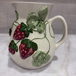 May include: A cream-colored ceramic pitcher featuring a raised, three-dimensional strawberry motif. The design includes glossy red strawberries with white seeds, vibrant green leaves, and thin vines wrapping around the body of the pitcher.