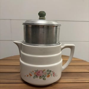May include: A white ceramic pitcher with a floral pattern featuring pink and orange flowers and green leaves. The pitcher has a cylindrical metal lid with a small, round, green-painted wooden knob handle on top and a side handle.