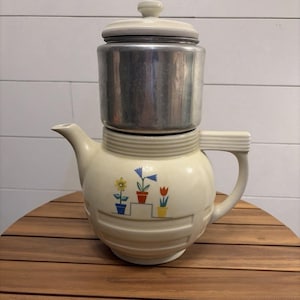 May include: A cream-colored ceramic teapot with a matching lid and a metallic silver-toned top section. The body features a graphic print of three potted flowers in blue, orange, and yellow pots arranged on a stepped platform design.