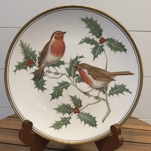 May include: A round white ceramic decorative plate featuring a detailed illustration of two robins perched on holly branches with green leaves and red berries. The plate has a thin black inner ring and a gold-colored rim along the outer edge.