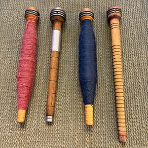 May include: Four wooden textile bobbins arranged vertically. The first is wrapped in red thread, the second is bare wood with a silver band, the third is wrapped in dark blue thread, and the fourth is a bare wooden bobbin with horizontal grooves.